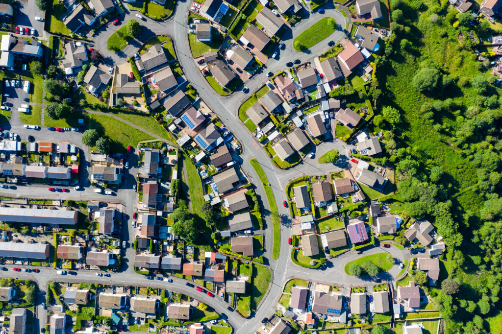 Aerial view of a suburban neighborhood with numerous houses, greenery, and curving streets, bordered by a forested area on the right side, showcasing potential for housing retrofits.